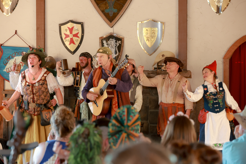 Nissa the Gnome holding a tiny mug on stage singing with a group of performers